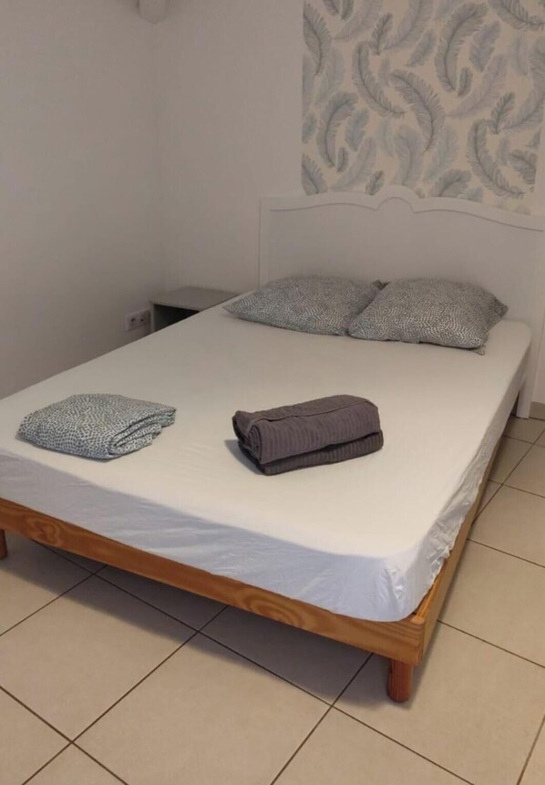 Un lit bien fait, avec un matelas blanc et deux oreillers, une couverture pliée et une serviette enroulée, sur un cadre de lit en bois, dans une pièce dont le sol est carrelé et un des murs est décoré d’un motif.