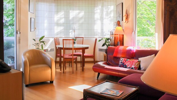 Ein Wohnzimmer mit Sofa, Sesseln, einem Esstisch und einem Fenster mit Blick ins Grüne.