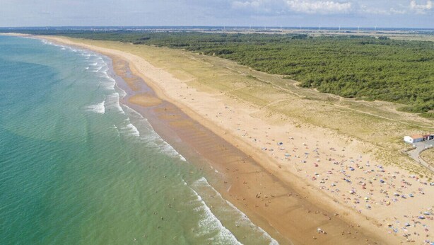Luftaufnahme eines Strandes mit einer weiten Sandfläche, einigen Strandsonnenschirmen und einem kleinen Gebäude am Wasser.