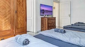 A bedroom with a large wooden wardrobe, a dresser, a bed with a blue mattress, and a wall-mounted TV displaying a scenic image.