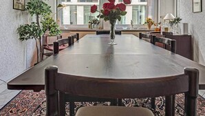 A long dining table with chairs, a vase of red roses, and a view of buildings through large windows.