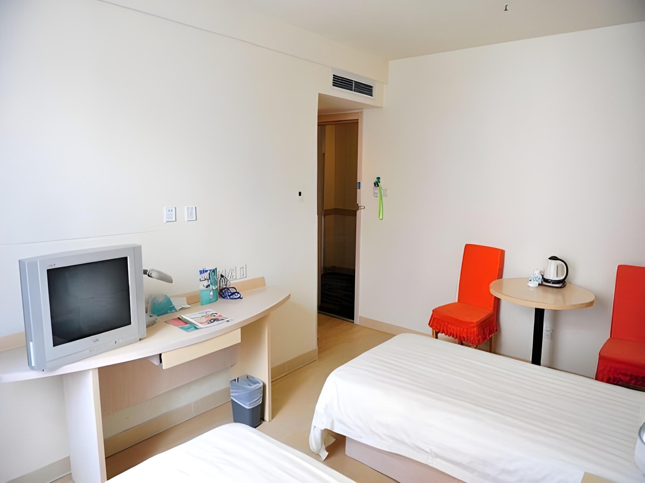 Quarto de hotel com televisão, secretária, duas camas, uma mesinha e duas cadeiras vermelhas.