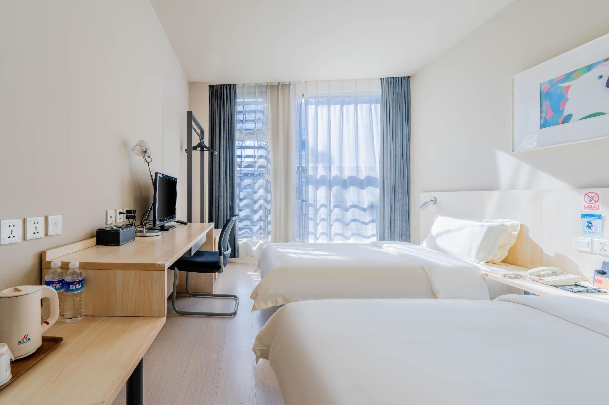 Une chambre d’hôtel avec deux lits, un bureau avec un ordinateur, une chaise, une télévision, une fenêtre avec des rideaux et un téléphone fixé au mur.