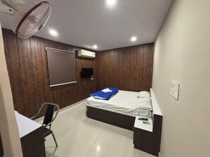 A small, modern bedroom with a bed, a desk, a chair, a TV, and a ceiling fan. - Hotel tuk tuk palace (Ranchi)