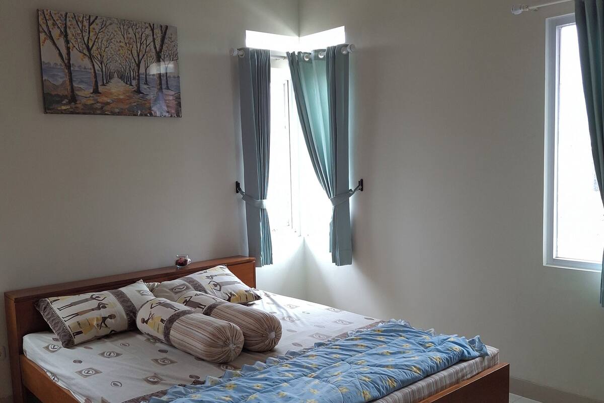 A bedroom with a wooden bed, a painting on the wall, and a window with curtains.