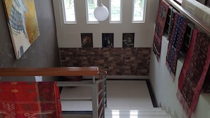 A staircase with a wooden handrail, a red patterned rug, and a wall with framed artwork and a patterned tapestry. - Dagope Homestay (Lembang)