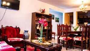 A living room with a TV, a red sofa, a wooden bookshelf, and a dining area with wooden chairs and a table. - Villa Flores (Mascota)