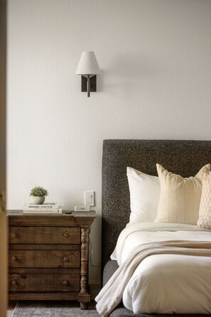 A bedroom with a wooden dresser, a bedside lamp, a bed with white bedding, and a textured wall. - Luxury Ranch: 10 Acres, Pool, Pickleball, Sauna, Party Barn (Dripping Springs)