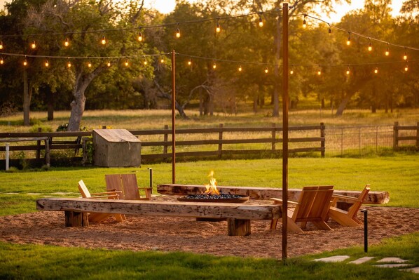An outdoor seating area with a fire pit, wooden benches, and string lights. - Deep Eddy Ranch: 10 Acres, Pool, Pickleball, Sauna (Dripping Springs)