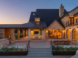 A two-story house with a stone facade, a covered patio with outdoor furniture, and a large window overlooking the yard. - Luxury Ranch: 10 Acres, Pool, Pickleball, Sauna, Party Barn (Dripping Springs)