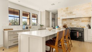 A modern kitchen with a large island, white cabinetry, and a stone fireplace. - Deep Eddy Ranch: 10 Acres, Pool, Pickleball, Sauna (Dripping Springs)