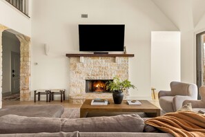 A living room with a stone fireplace, a flat-screen TV, a sofa, a coffee table, and a potted plant. - Deep Eddy Ranch: 10 Acres, Pool, Pickleball, Sauna (Dripping Springs)