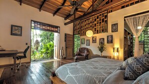 Ein geräumiges Schlafzimmer mit einem großen Bett, einem Schreibtisch und einem Sessel, einem Deckenventilator und Blick auf üppiges Grün durch die Fenster.