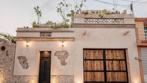A building with a large wooden door, a small entrance with a metal gate, and a stone wall with a plant-covered roof. - Casa Ek: House•Safe•Fast WiFi•Pool•Walkable  area (Mérida)