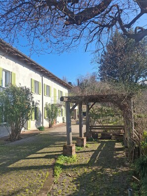 A rustic building with a tiled roof, green shutters, and a garden area with a pergola. - Former wine estate with breathtaking panoramic views! (Canelli)