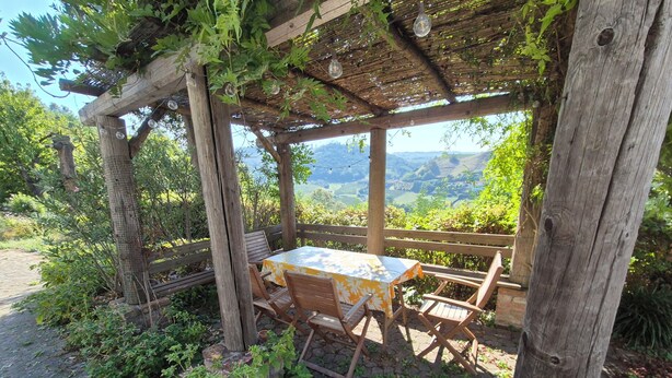 A wooden pergola with a table and chairs underneath, surrounded by greenery and a view of the countryside. - Former wine estate with breathtaking panoramic views! (Canelli)