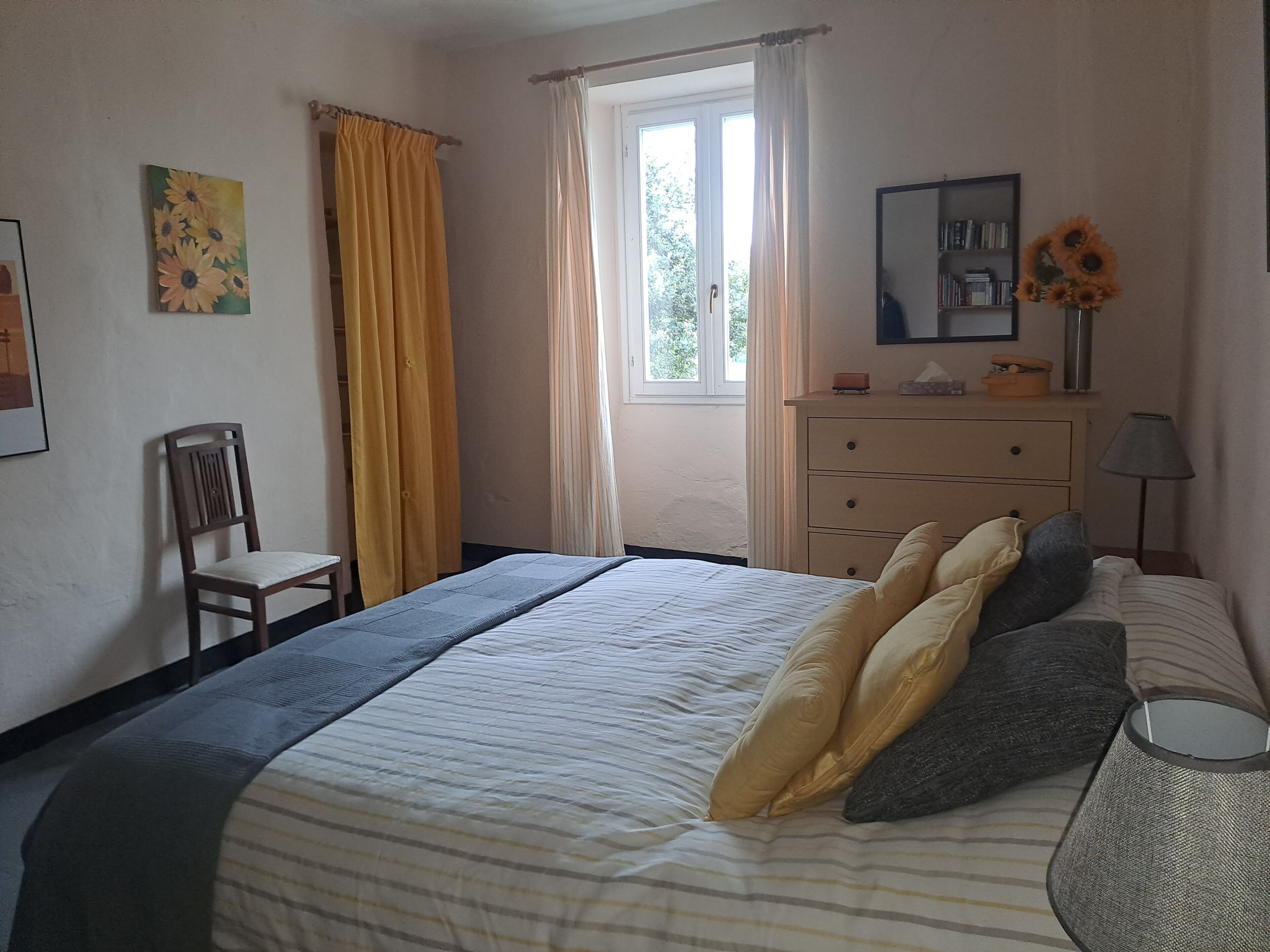 Un dormitorio con cama, una silla, cómoda, ventana con cortinas, un cuadro y un jarrón con flores.
