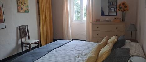 Un dormitorio con cama, una silla, cómoda, ventana con cortinas, un cuadro y un jarrón con flores.