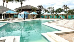 A swimming pool with lounge chairs, palm trees, and thatched-roof structures.