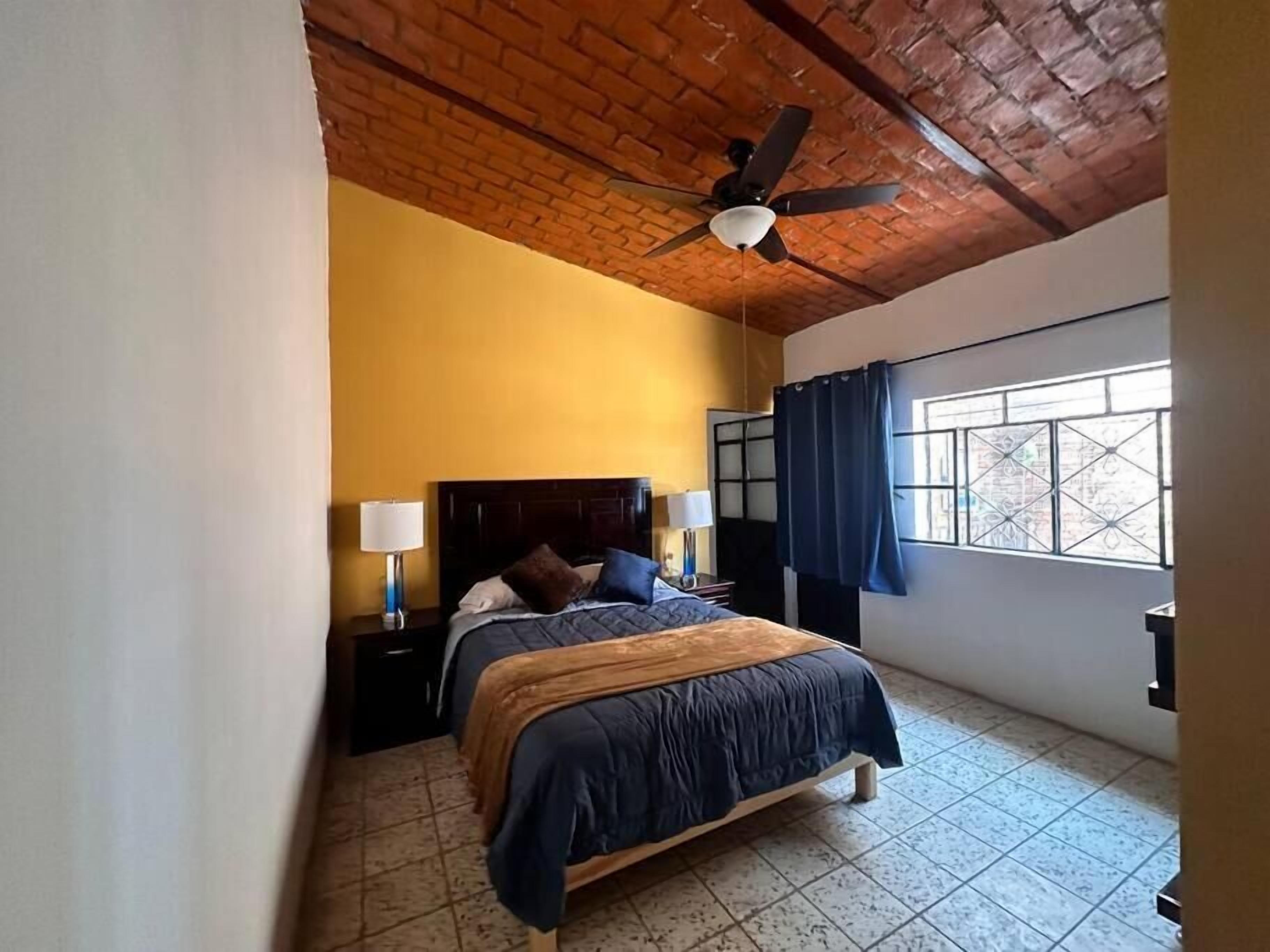 A bedroom with a wooden ceiling, a bed with a dark blue bedspread, two bedside lamps, and a window with blue curtains.