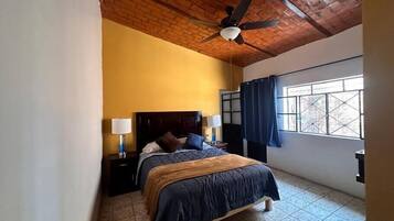 A bedroom with a wooden ceiling, a bed with a dark blue bedspread, two bedside lamps, and a window with blue curtains.