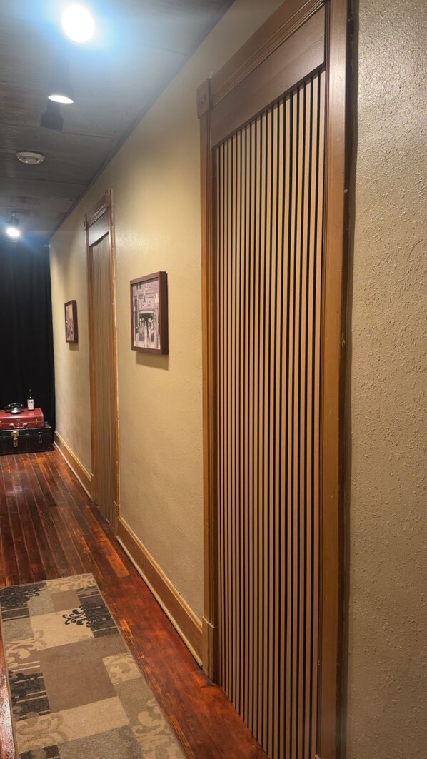 A hallway with wooden flooring, a patterned rug, a closed door with vertical blinds, and a framed picture on the wall.
