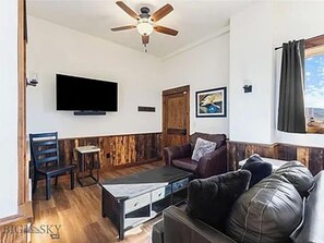 A living room with a television, a ceiling fan, a wooden accent wall, a sofa, and a window with curtains.