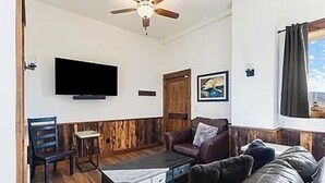 Una sala de estar con ventilador de techo, televisor de pantalla plana, sofá, mesa de centro y ventana con cortinas.
