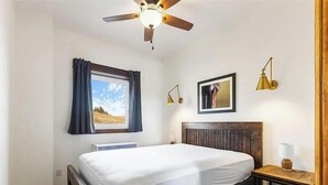 Un dormitorio con cama, ventilador de techo, ventana con cortinas, mesita de noche con lámpara y un cuadro en la pared.