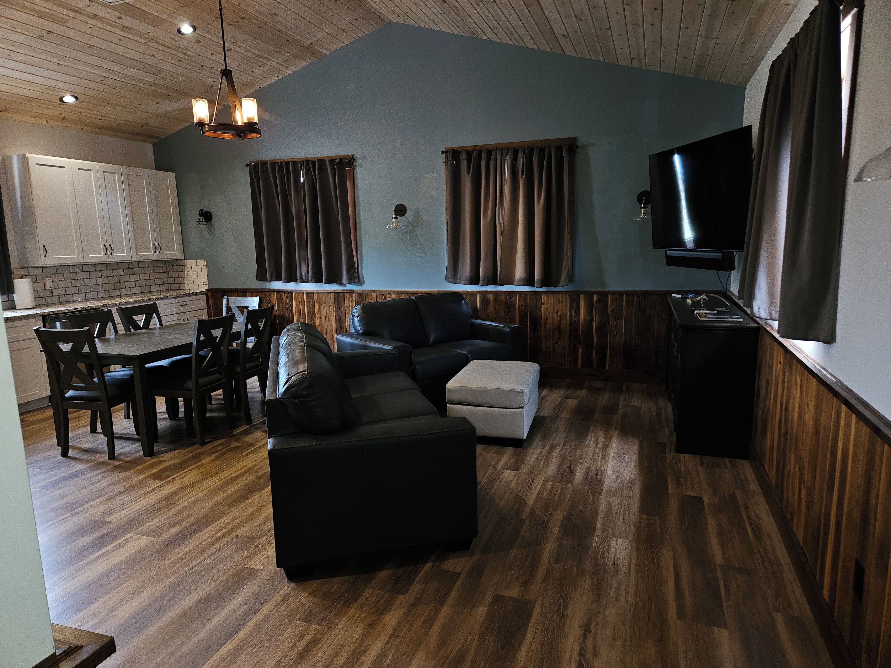 Un salon moderne avec un canapé d’angle, un coin repas avec une table et des chaises, une cuisine avec des meubles blancs et un téléviseur à écran plat fixé au mur.