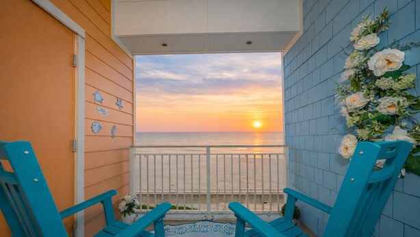 A balcony with two turquoise chairs, a view of the sunset over the ocean, and a decorative wall with flowers. - New! Oceanfront / Pool / Boat / Surf / Kayak, Swim (Virginia Beach)