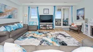 A living room with a sofa, TV, and a view to the outside. - New! Oceanfront / Pool / Boat / Surf / Kayak, Swim (Virginia Beach)