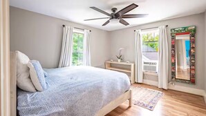 A bedroom with a bed, a ceiling fan, a window with curtains, a small bedside table, and a mirror with a decorative frame. - Sunny House, By The Beach, Minutes From Portland (South Portland)