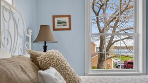Ein Schlafzimmer mit großem Fenster, einem Bett mit Kissen, einer Lampe und einem gerahmten Bild an der Wand.