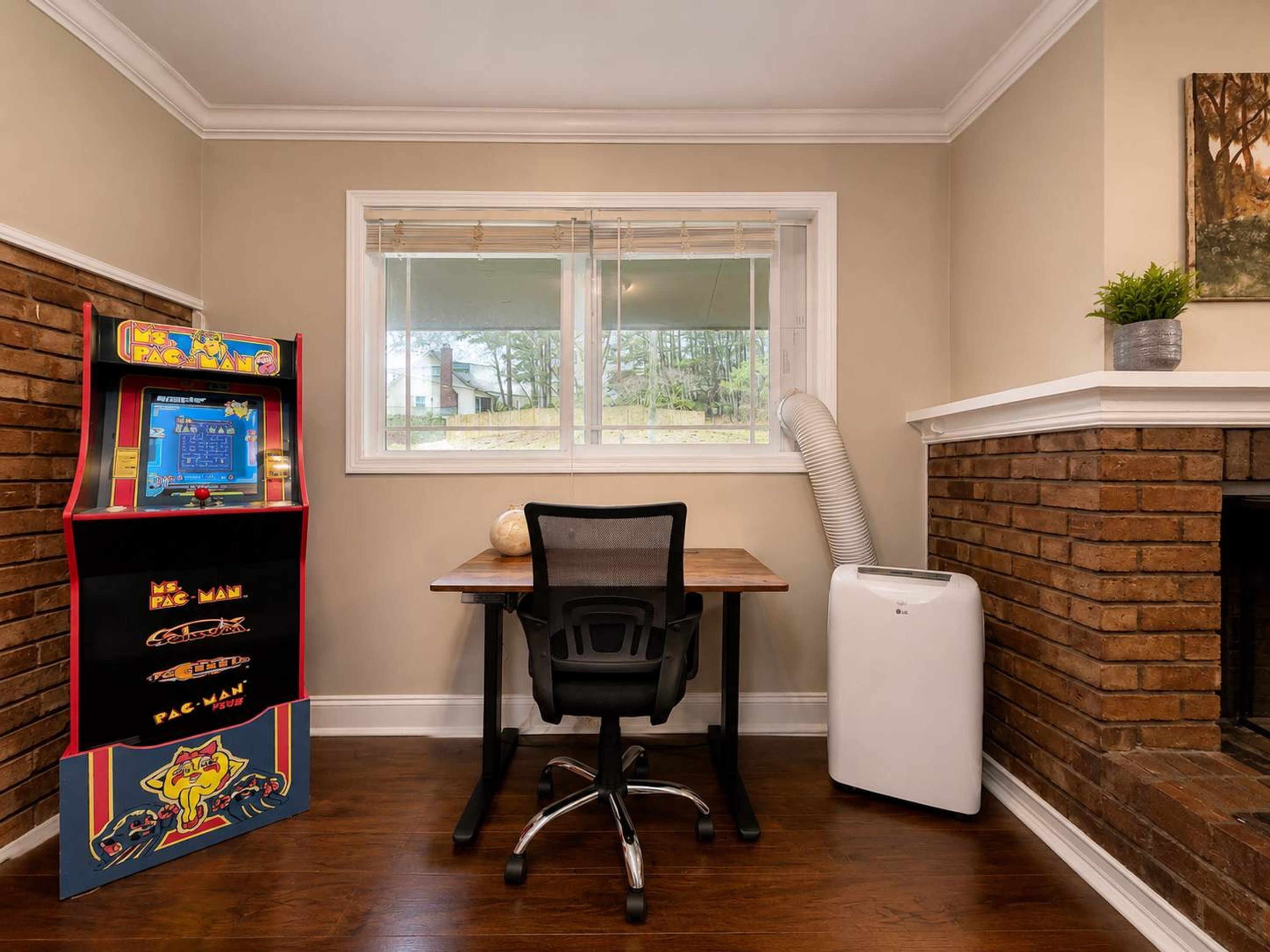 A room with a wooden desk, a black office chair, a window with blinds, a fireplace, and a Pac-Man arcade machine.