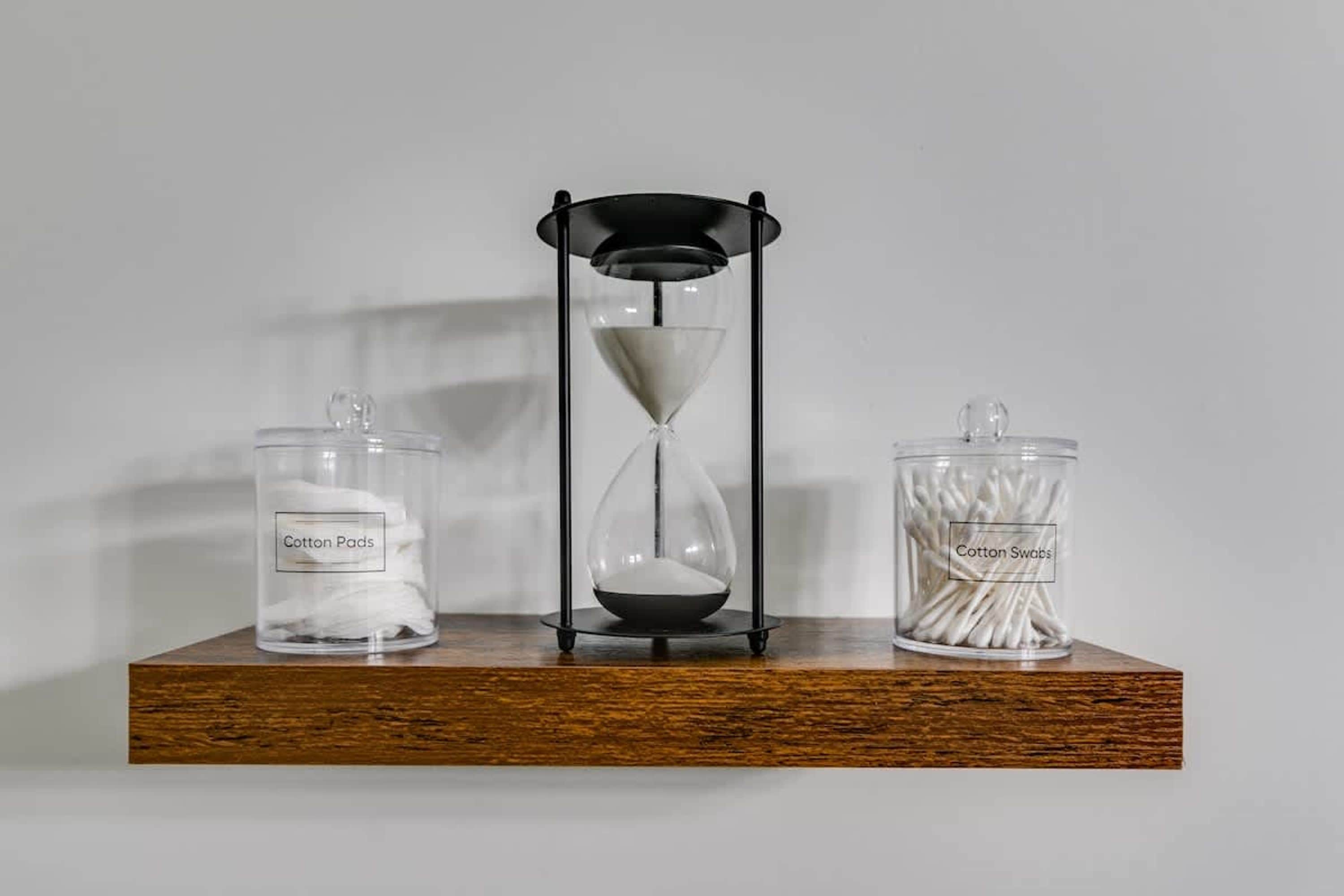 A wooden shelf with three glass containers labeled 'Cotton Pads,' 'Cotton Swabs,' and an hourglass.