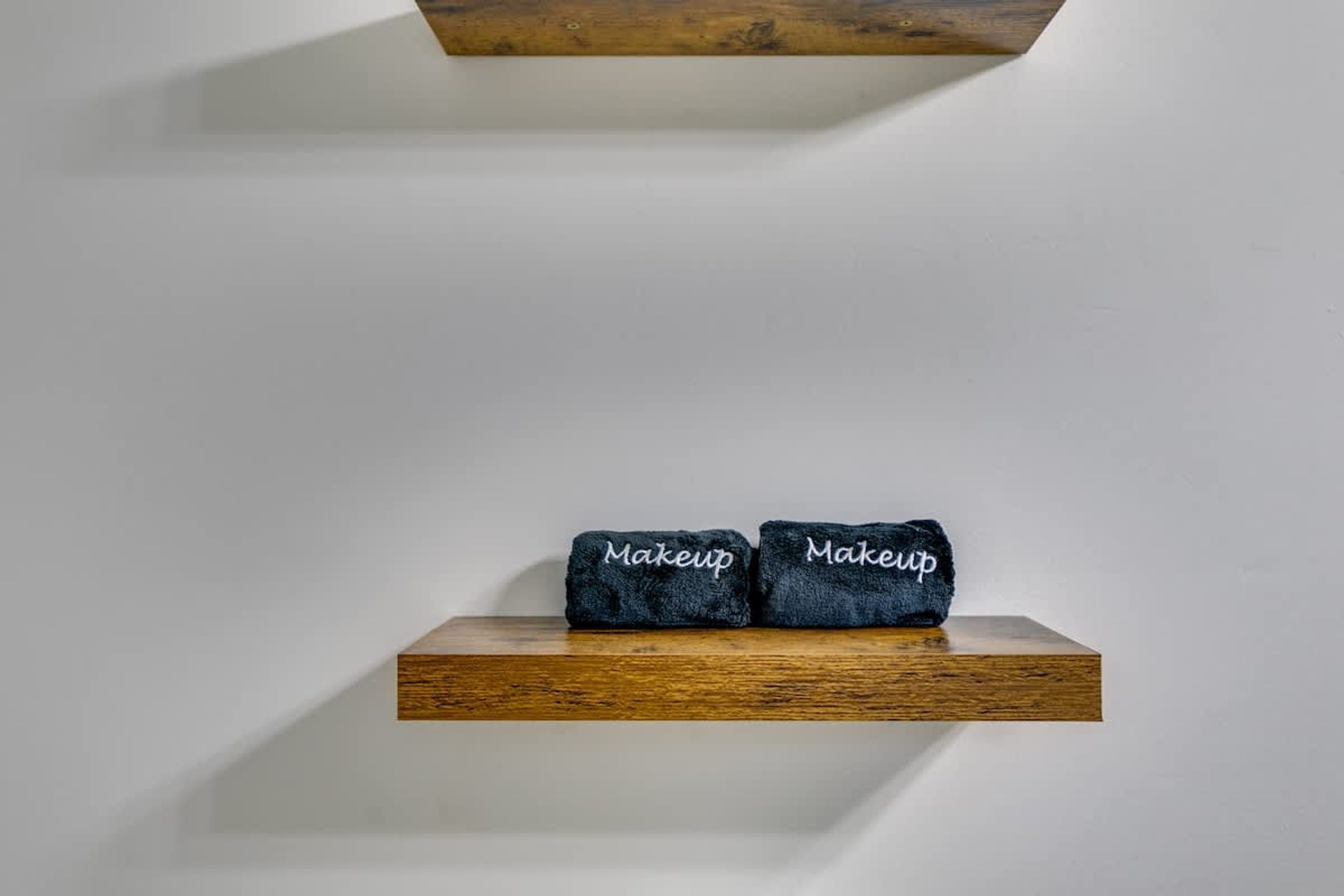 Un estante de madera con dos toallas azul oscuro etiquetadas 'Makeup' frente a una pared blanca lisa.