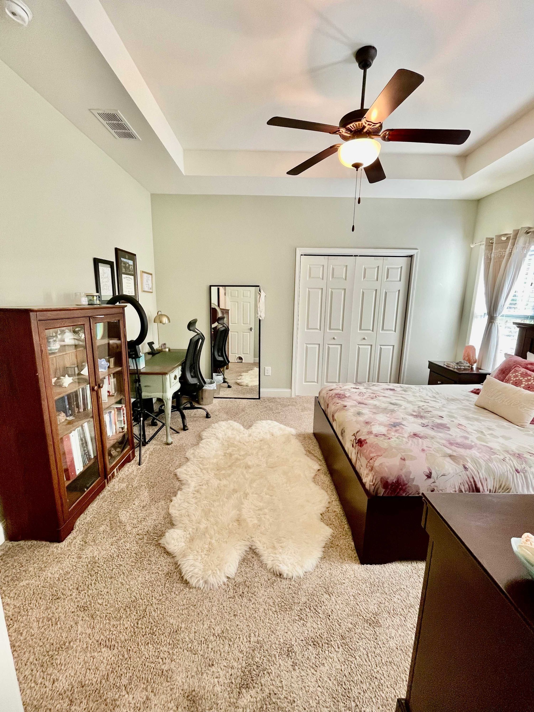 A bedroom with a ceiling fan, a bed with a floral bedspread, a desk with a chair, a mirror, and a wardrobe.