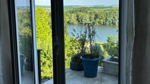 Vista attraverso una porta di vetro su un balcone con piante in vaso, un pavimento in cemento e un lago sullo sfondo.