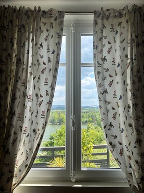 Une fenêtre ornée de rideaux à motifs floraux, donnant sur un paysage verdoyant et un lac.