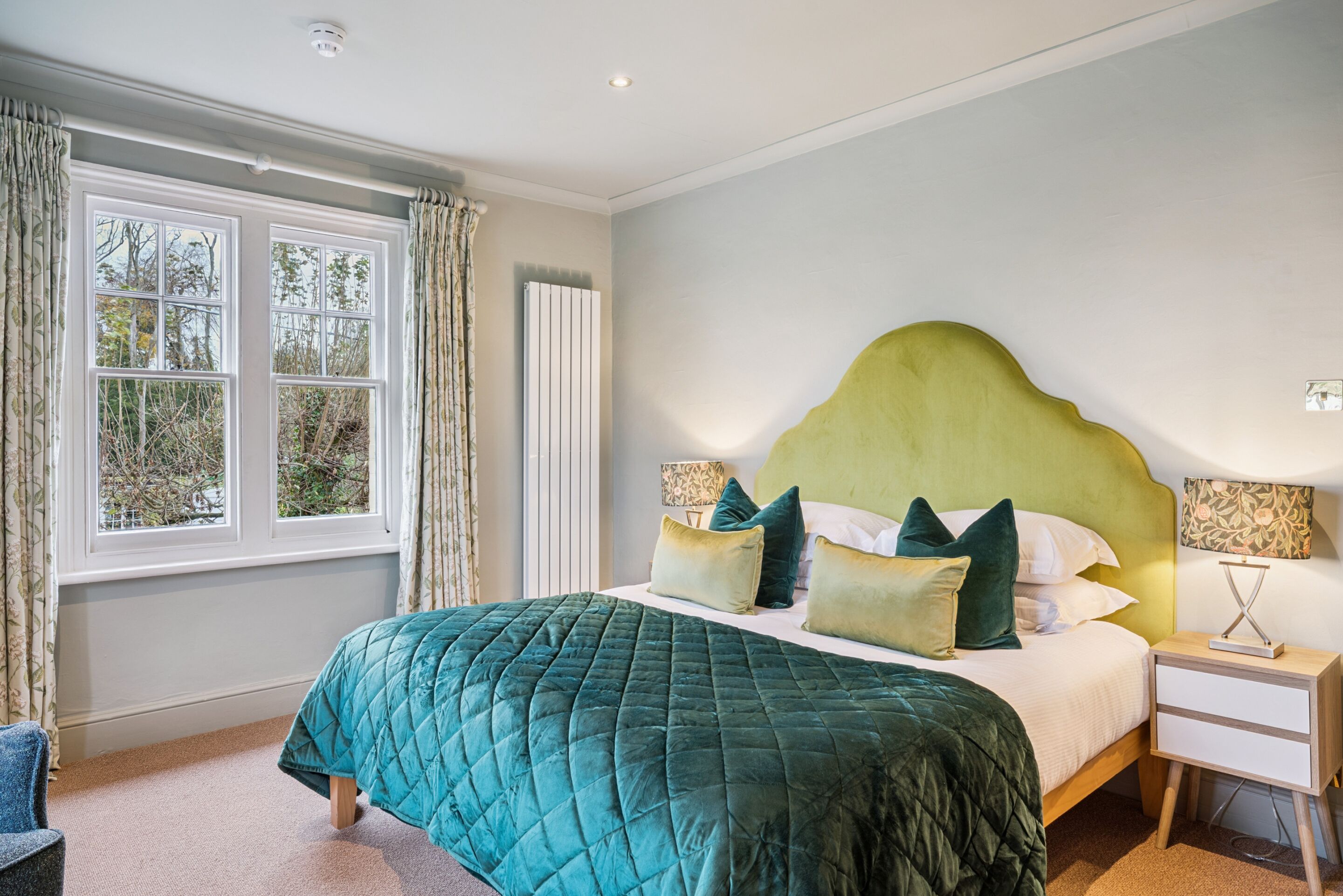 A bedroom with a large bed, a nightstand, a lamp, a window with curtains, and a radiator.