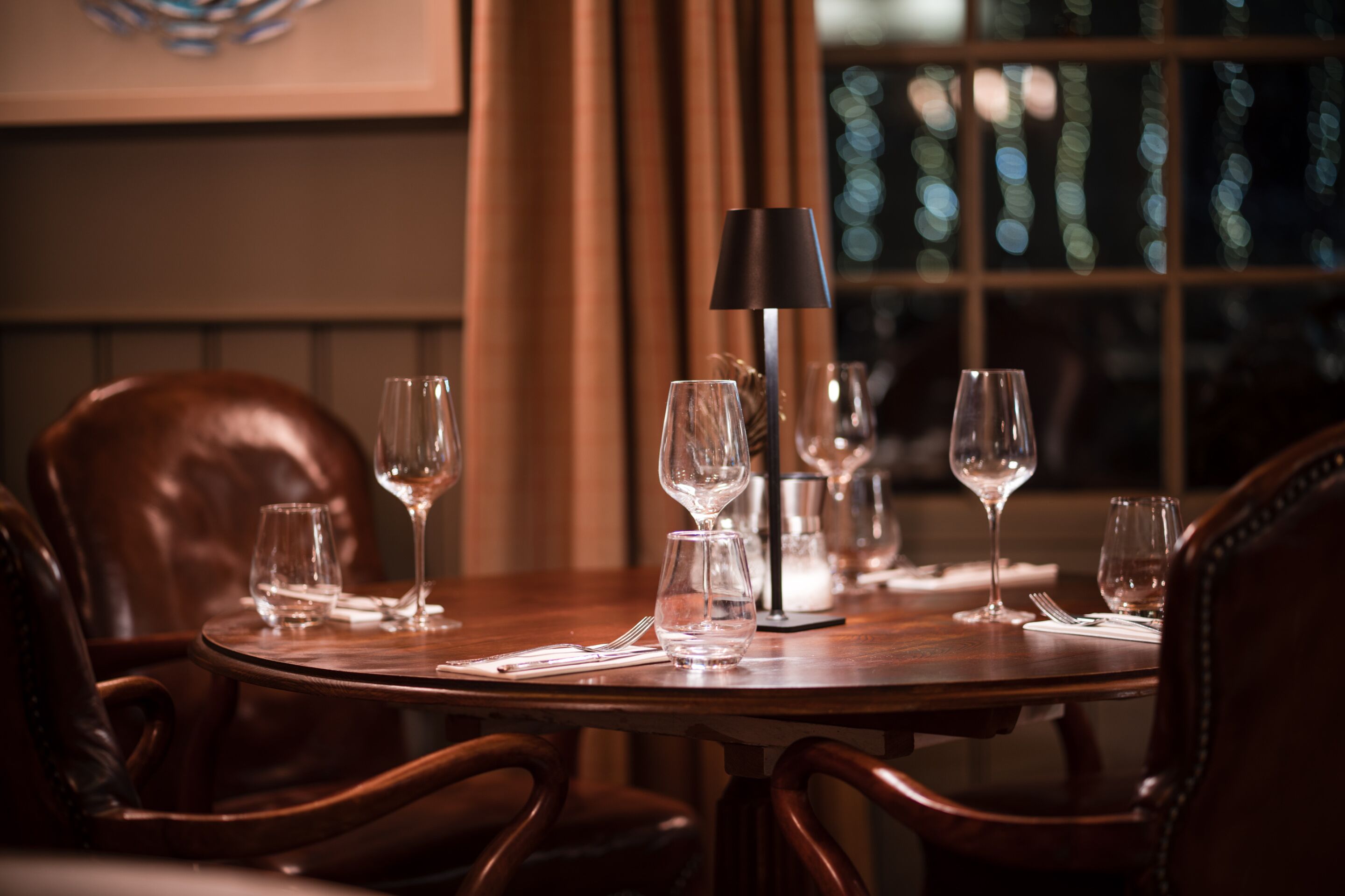 Une table à manger, agrémentée de verres à vin et d’une lampe, entourée de fauteuils en cuir.