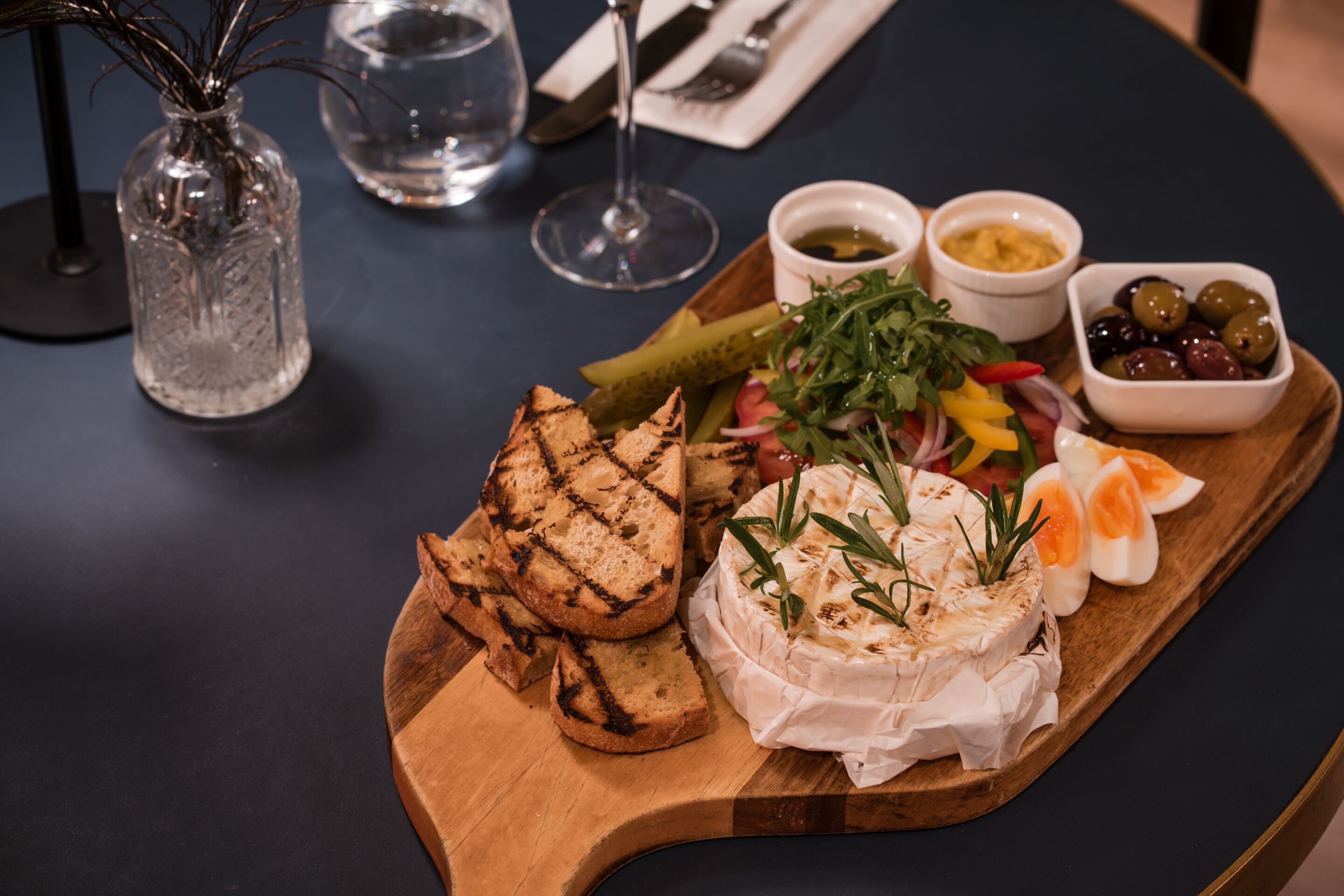 A wooden board with grilled bread, a cheese wheel, and a salad on a table.