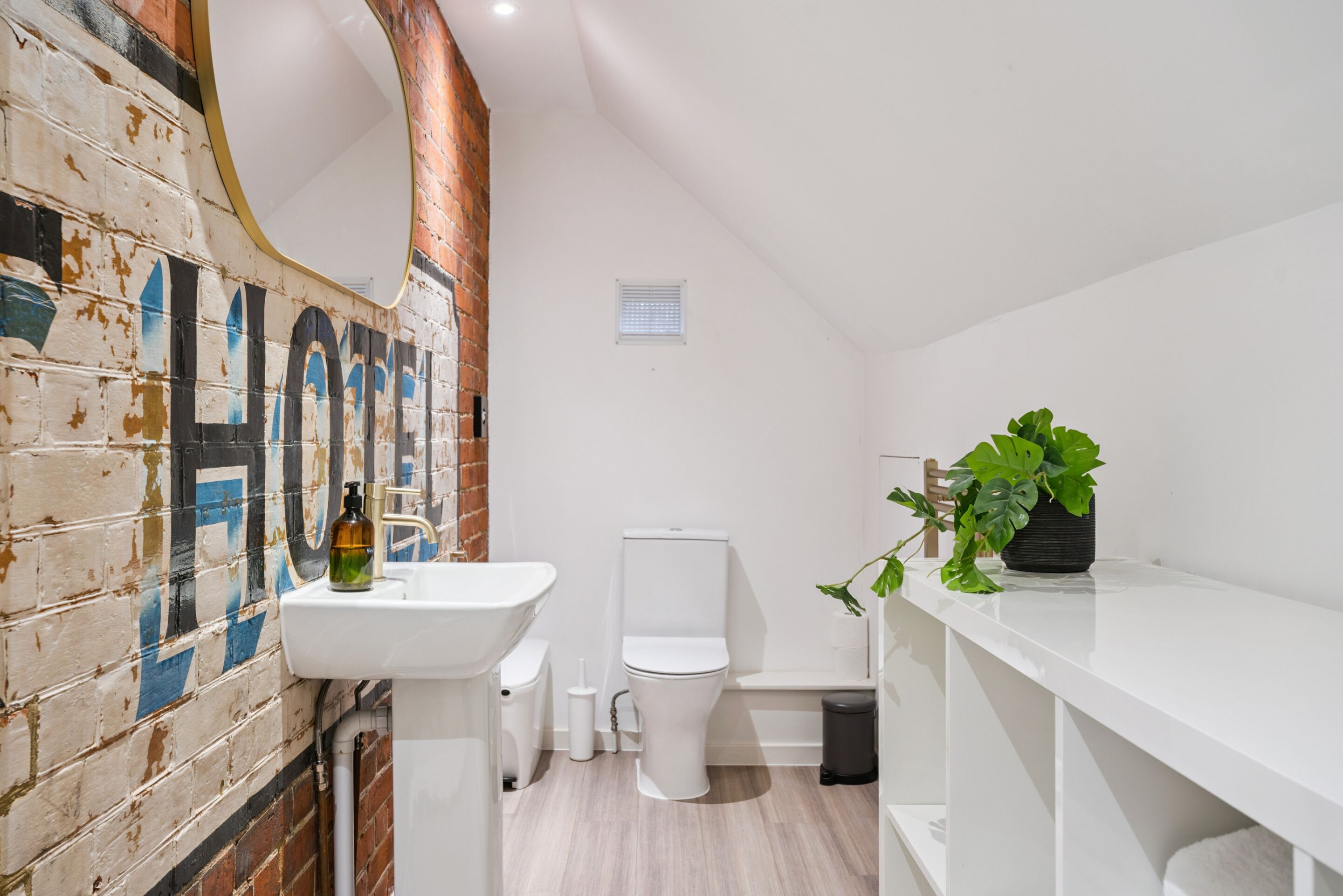 A bathroom with a white sink, toilet, and a mirror. There is a plant on the right side and a brick wall with a mural on the left.