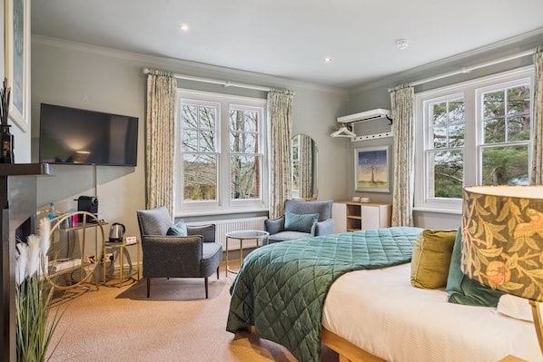 A bedroom with a bed, a chair, a television, a heater, and large windows. - The Flower Pot Hotel (Henley-on-Thames)