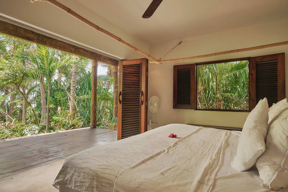 A bedroom with a bed, wooden shutters, a fan, and a view of tropical vegetation.