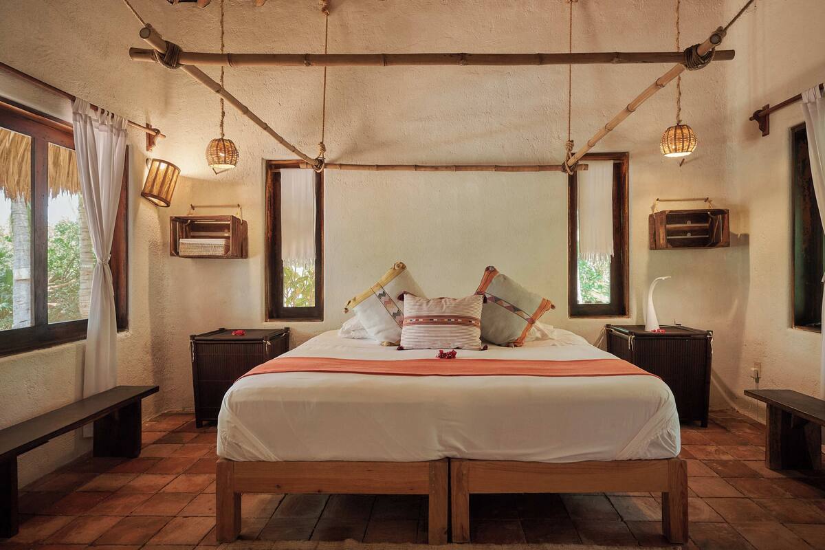A bedroom with a wooden bed, two bedside tables, a bench, a window with curtains, and a hanging light fixture.