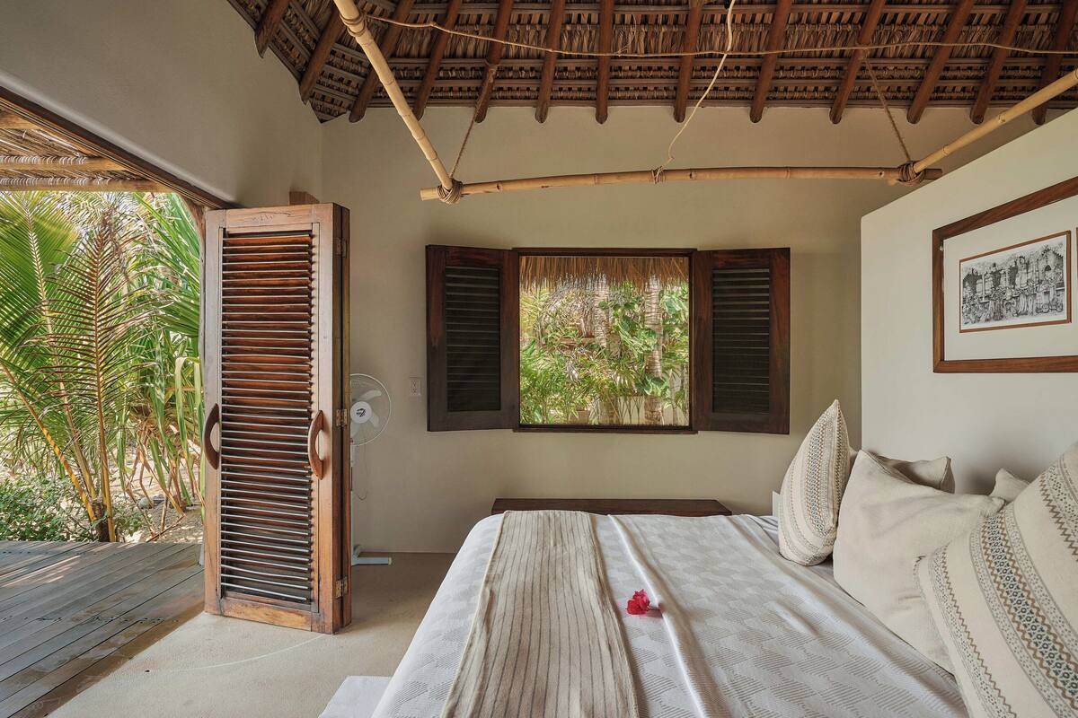 A bedroom with a bed, wooden shutters, a fan, and a window with a view of greenery.