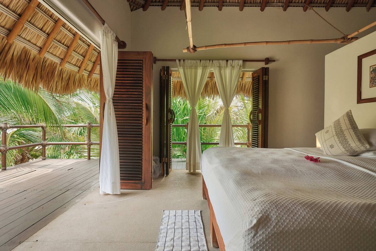 A room with a bed, a window with curtains, a wooden ceiling, and a view of greenery outside.
