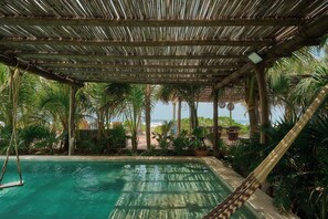 Zona de piscina con techo de paja, rodeada de palmeras y vista a la playa.